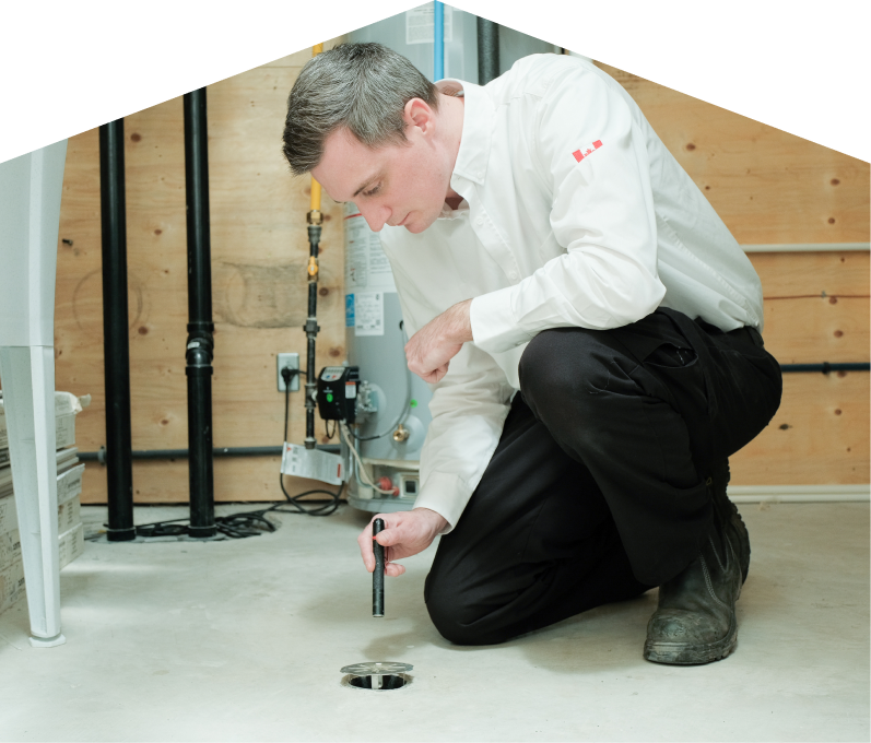 Limcan plumbing specialist inspecting a basement floor drain with a flashlight during a drain cleaning service, in a utility room with visible pipes, water heater, and wood panel walls. Limcan plumbing specialist inspecting a basement floor drain with a flashlight during a drain cleaning service, in a utility room with visible pipes, water heater, and wood panel walls.